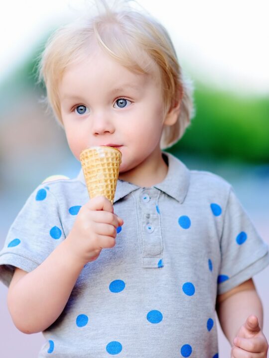 Boy in a gray shirt with blue dots eating an ice cream cone.