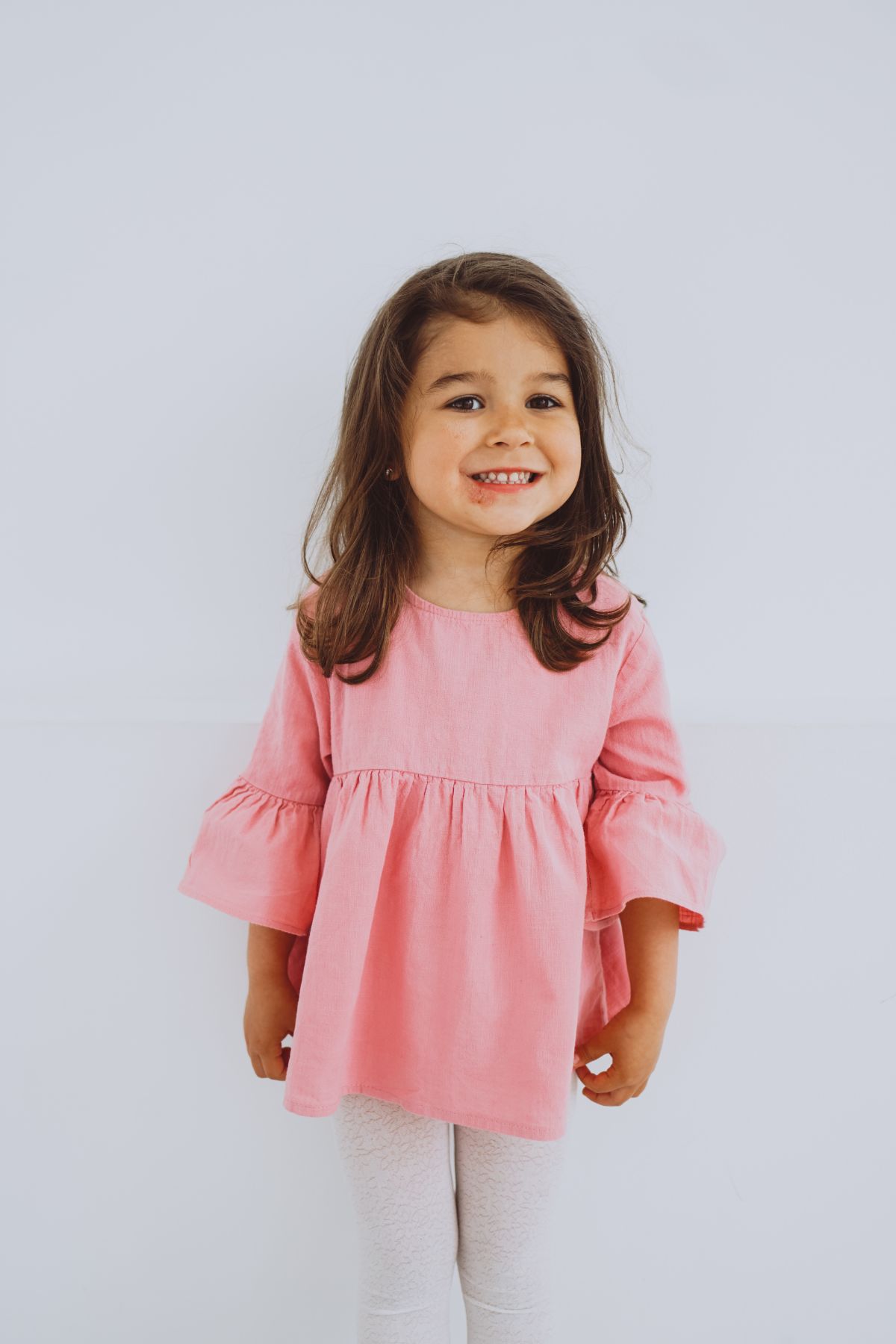 Little brunette girl in pink shirt smiling and standing by gray background.