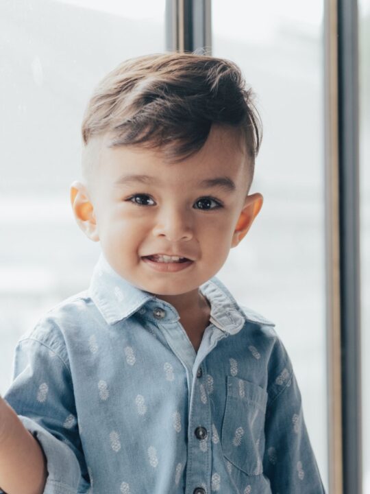 Little boy with brown hair and button down shirt standing by window and smiling.