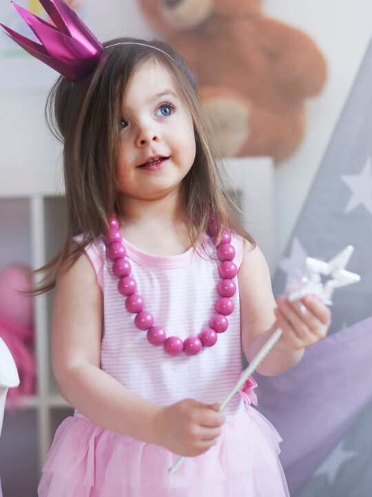 Little girl in pink dress, crown, and necklace holding star wand standing in playroom.