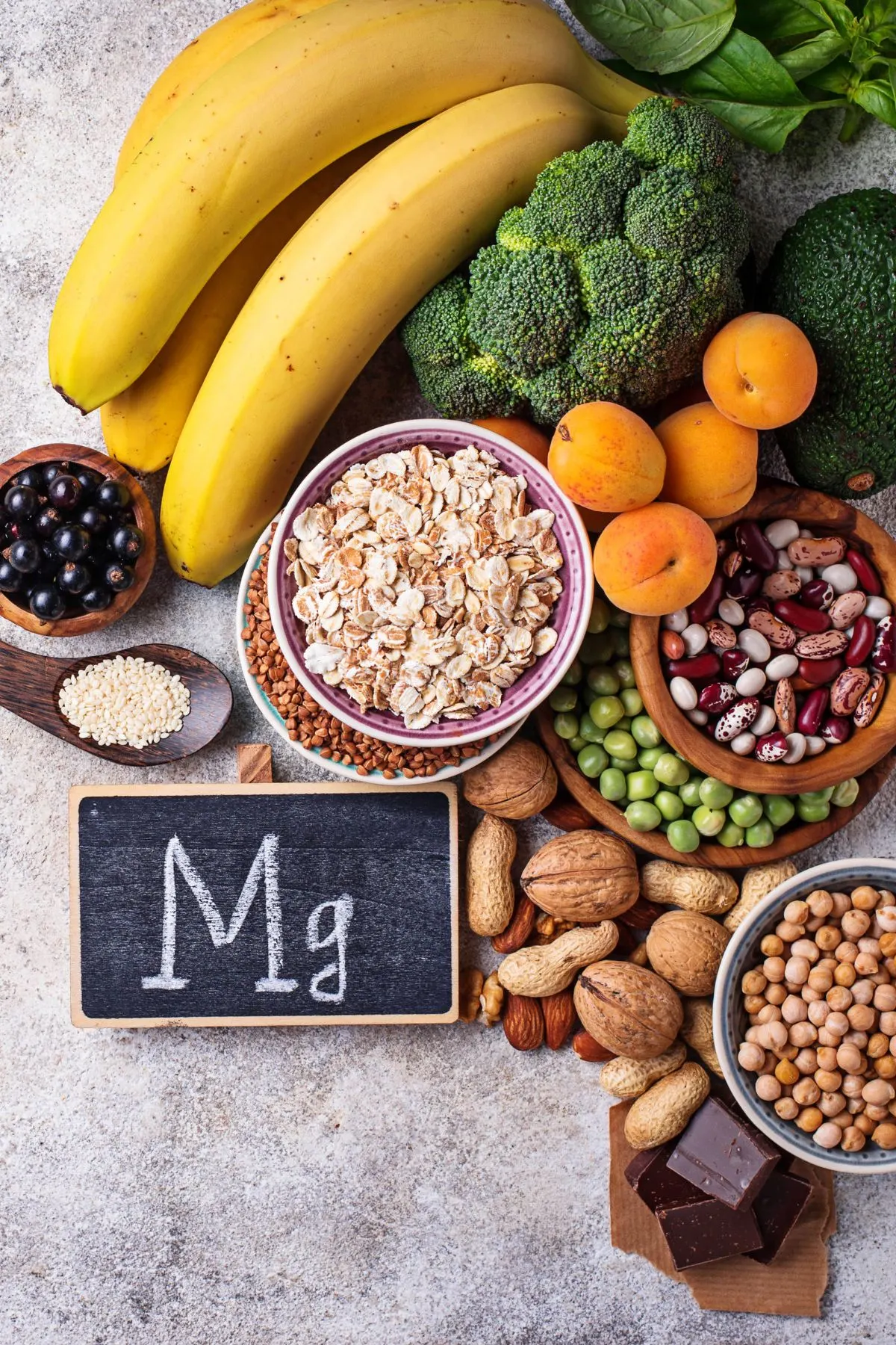 The letters "Mg" on a chalkboard surrounded by healthy fruits, vegetables, nuts, and seeds.
