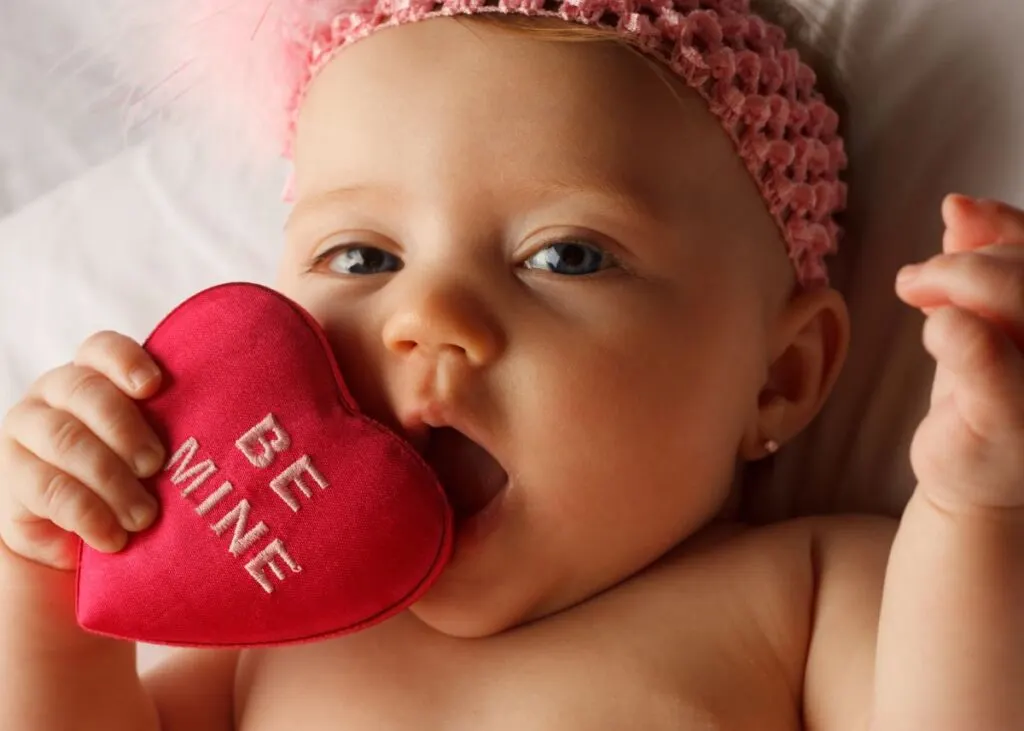 Baby girl with pink headband puts felt heart that says 'Be Mine' to her mouth.