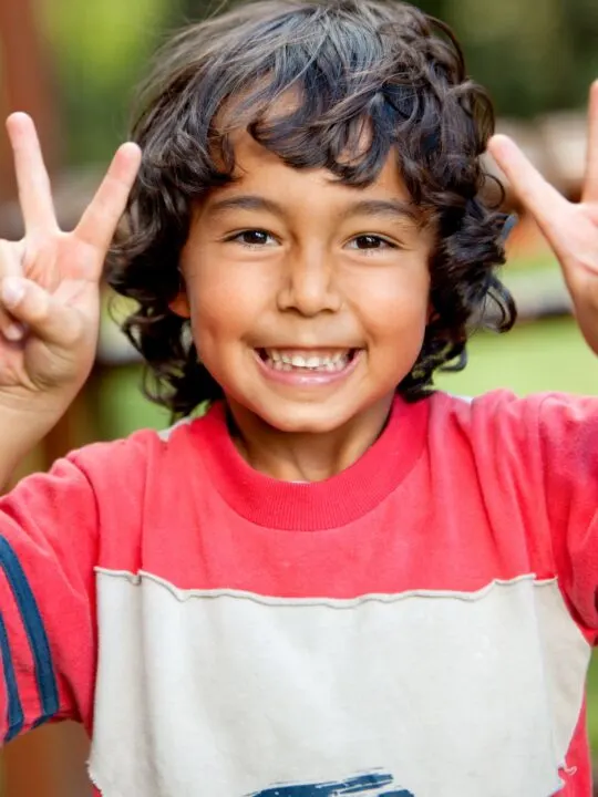A boy with red and gray shirt holds up two peace signs.