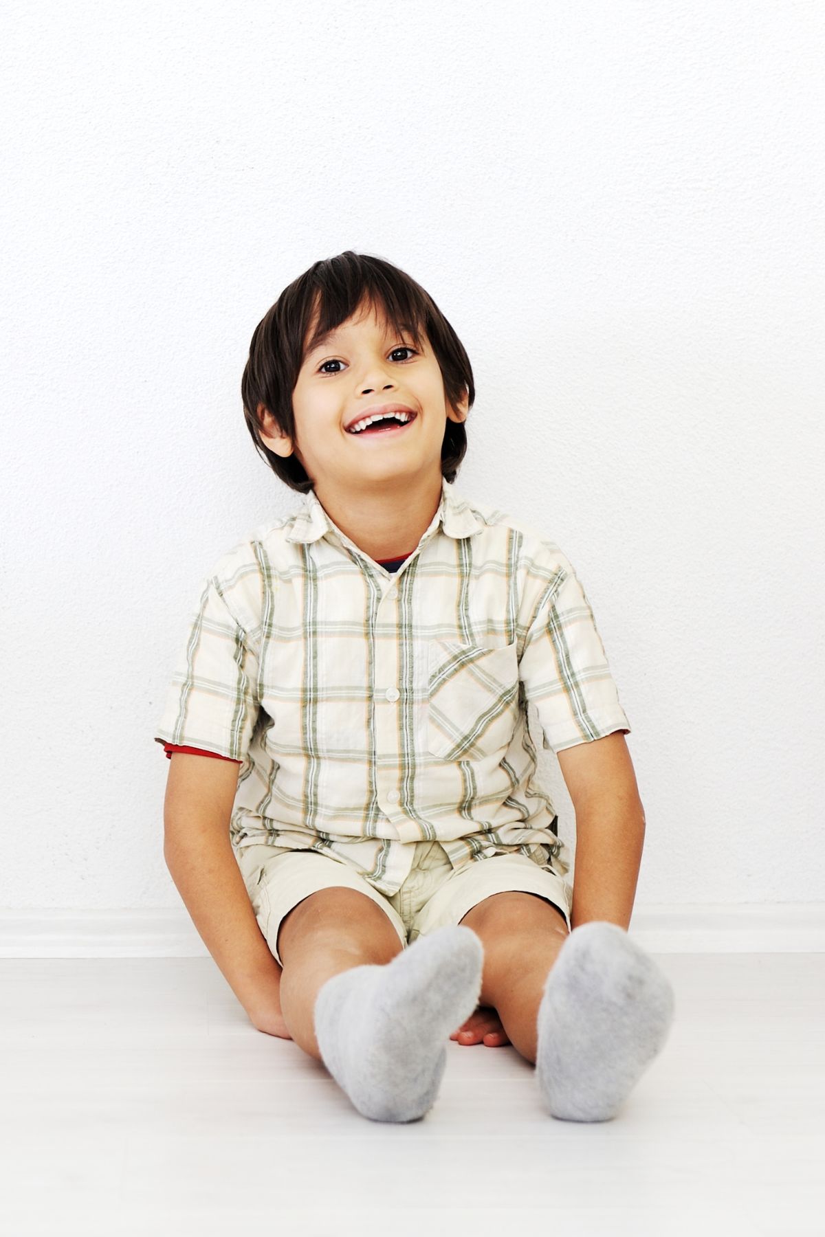 Smiling boy in a plaid shirt sits on the floor with his back to the wall.