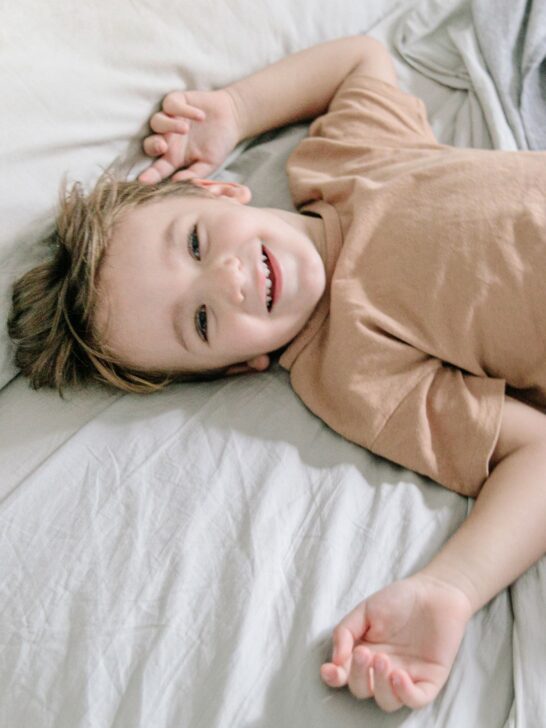 A smiling boy in a brown shirt laying on a bed with a white sheet.