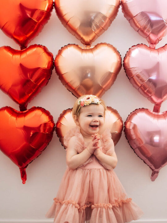 Baby girl wearing pink dress claps hands in front of heart balloon backdrop.