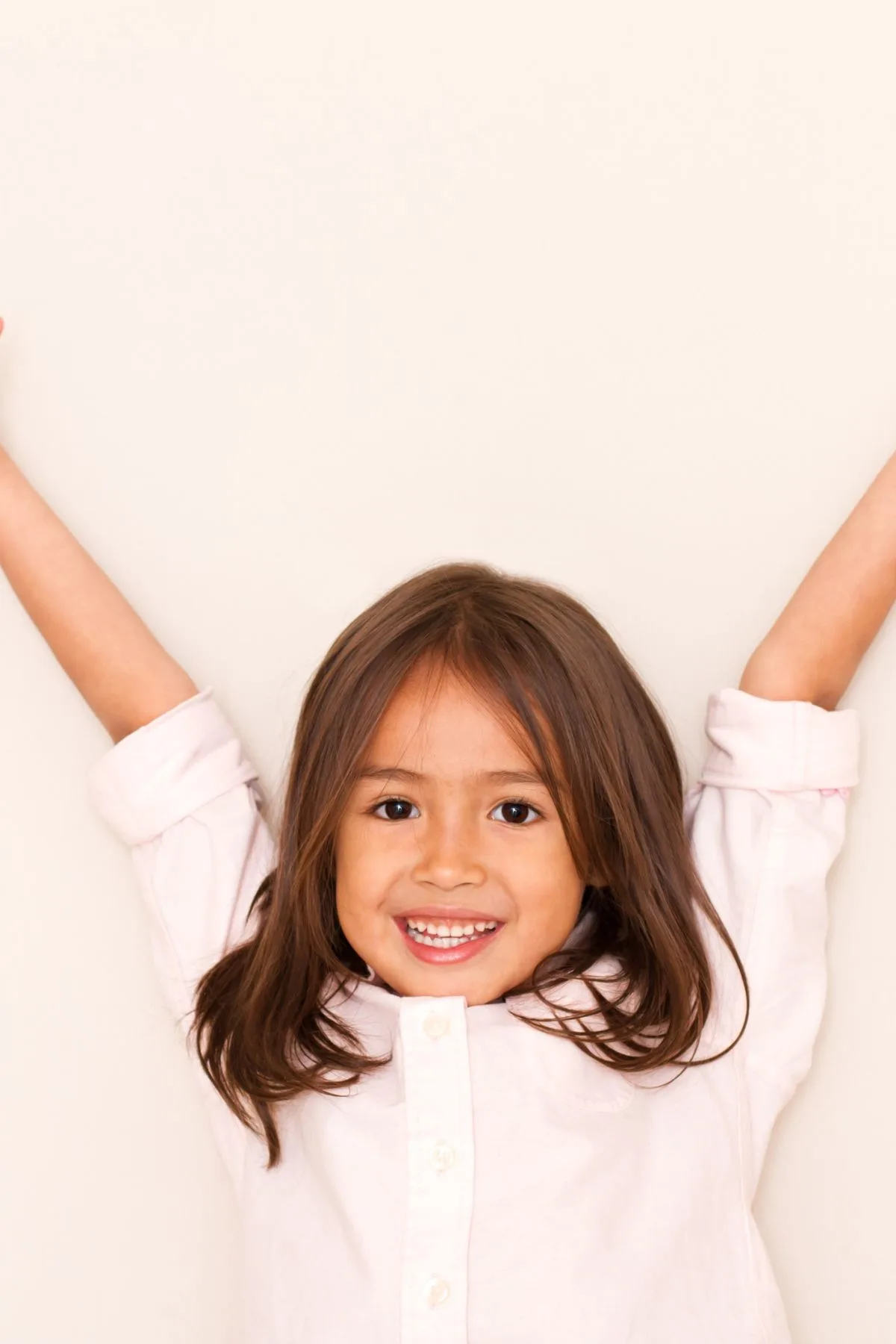 A girl stands in front of a cream background with arms raised in excitement.