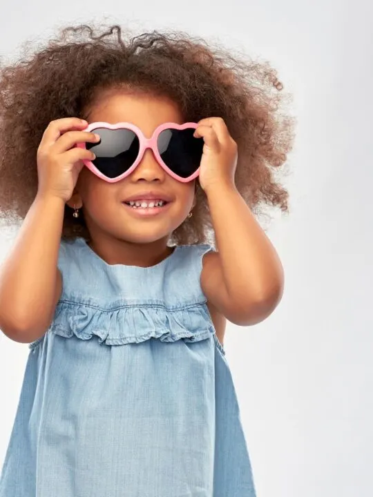 Girl in a denim dress holds pink heart-shaped sunglasses to her face.