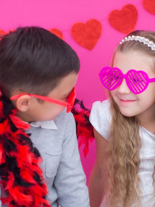 Kids wearing heart-shaped glasses in front of a heart banner and pink background.