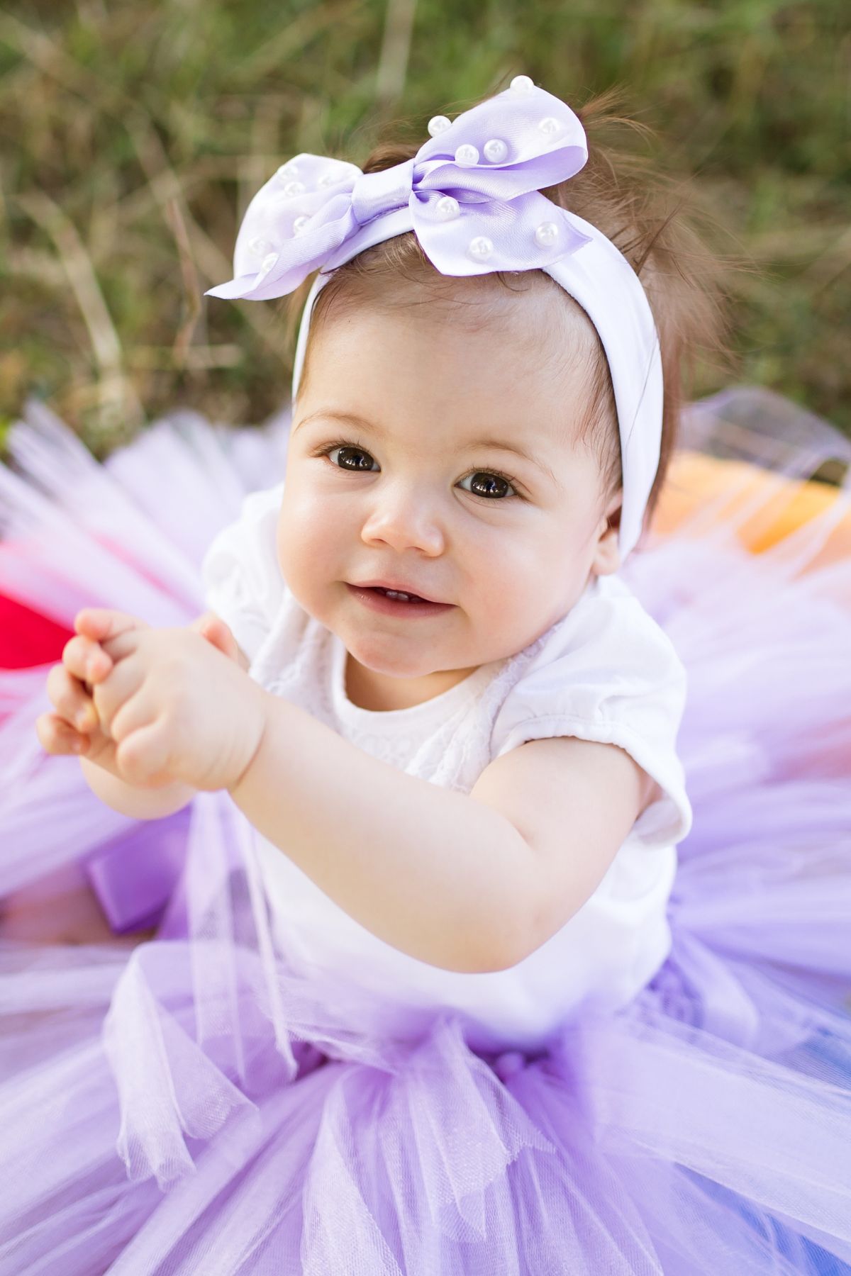 Baby girl in purple dress and headband with bow sitting outside.