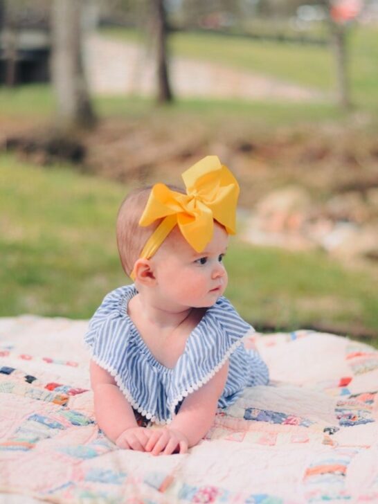 Baby girl with yellow large yellow bow on a blanket outside.