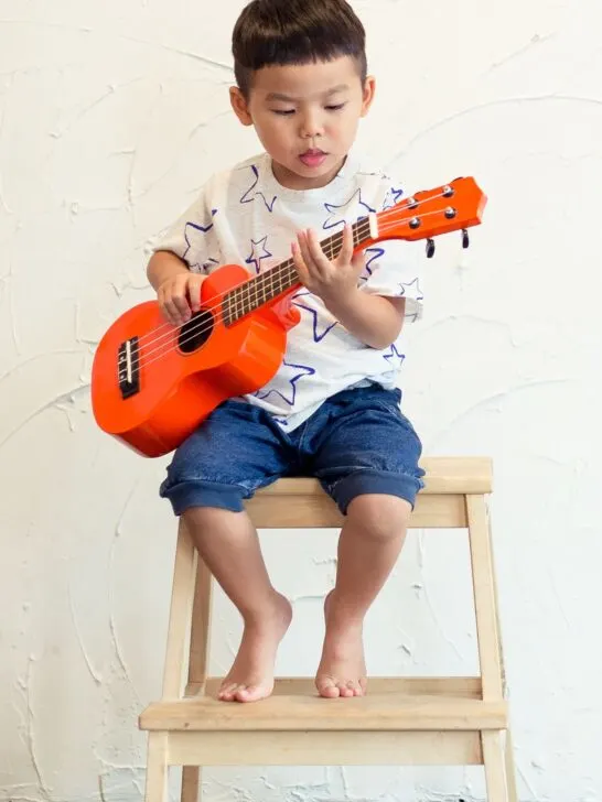 A toddler boy plays a red guitar while sitting on a wooden stool.