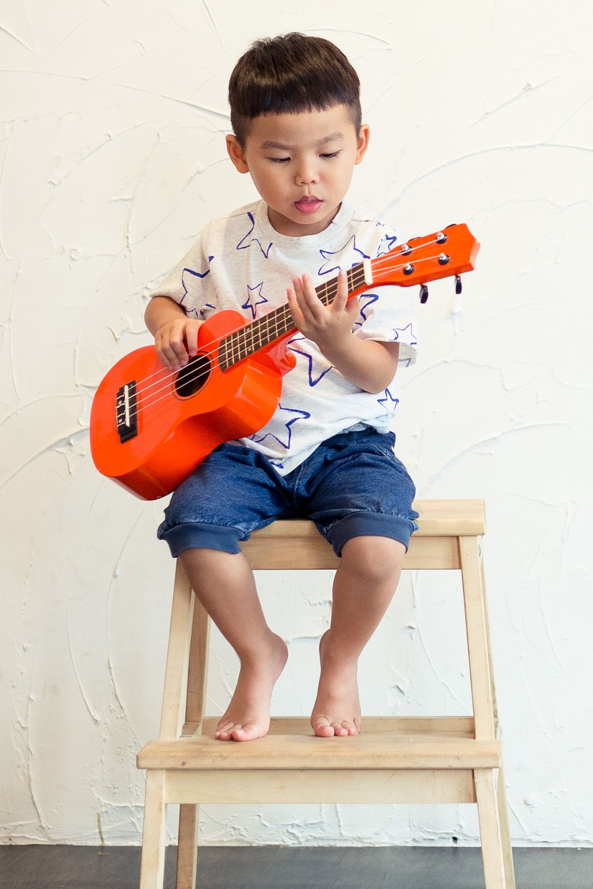 A toddler boy plays a red guitar while sitting on a wooden stool.