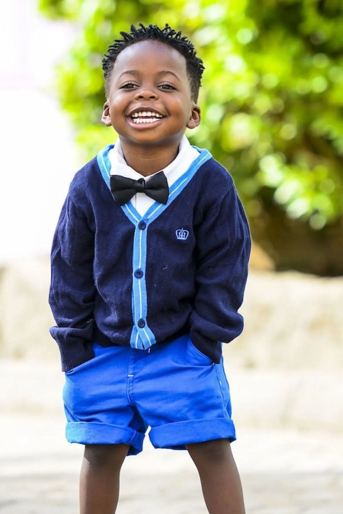 Smiling boy in a blue sweater, shorts, and bowtie, standing outside.