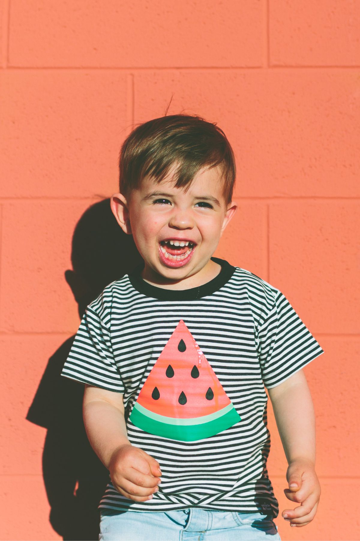 A boy with a watermelon shirt on laughing stands in front of an orange brick wall.