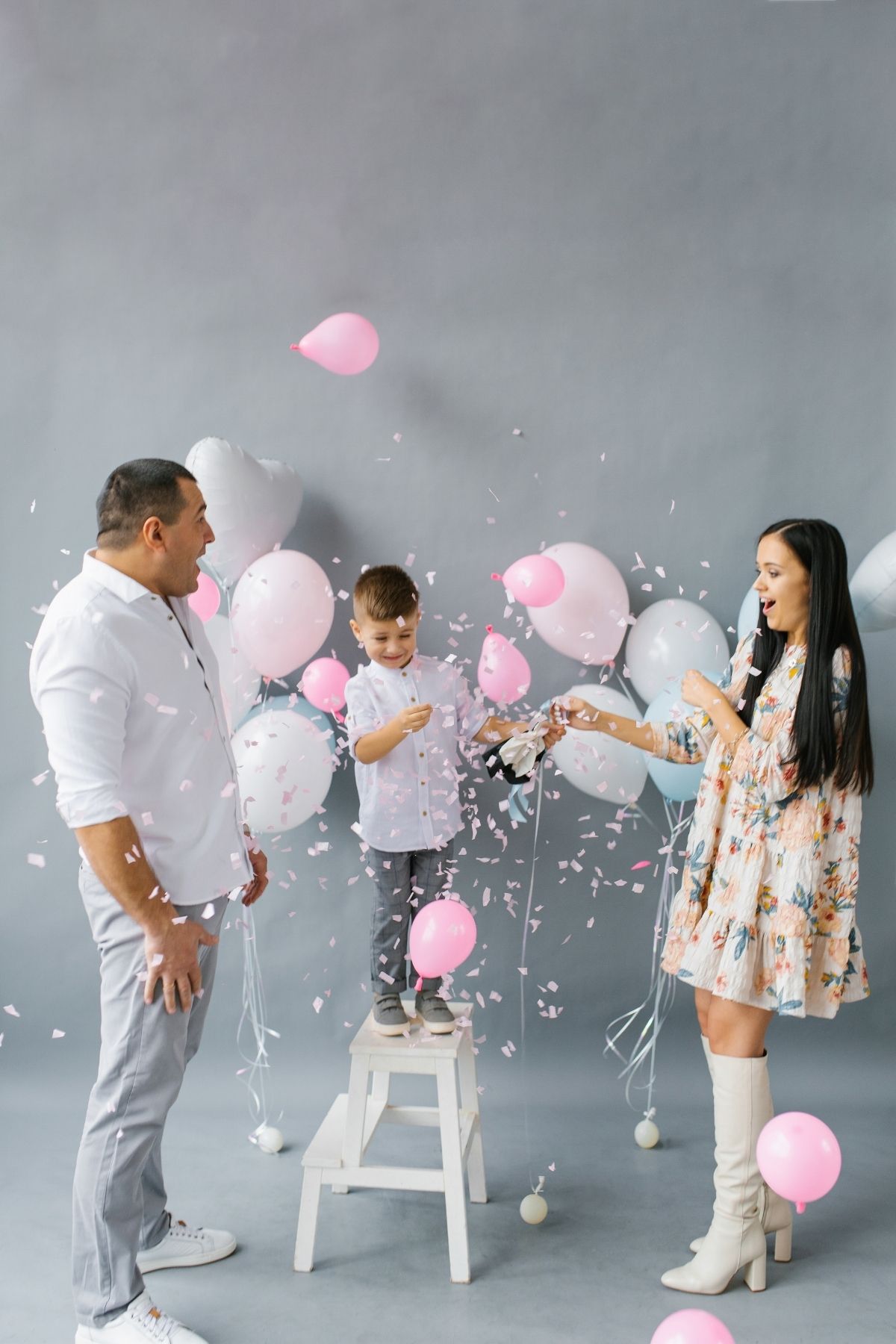 Man and woman smile with excitement next to boy on stool while pink confetti and balloons fall to ground.