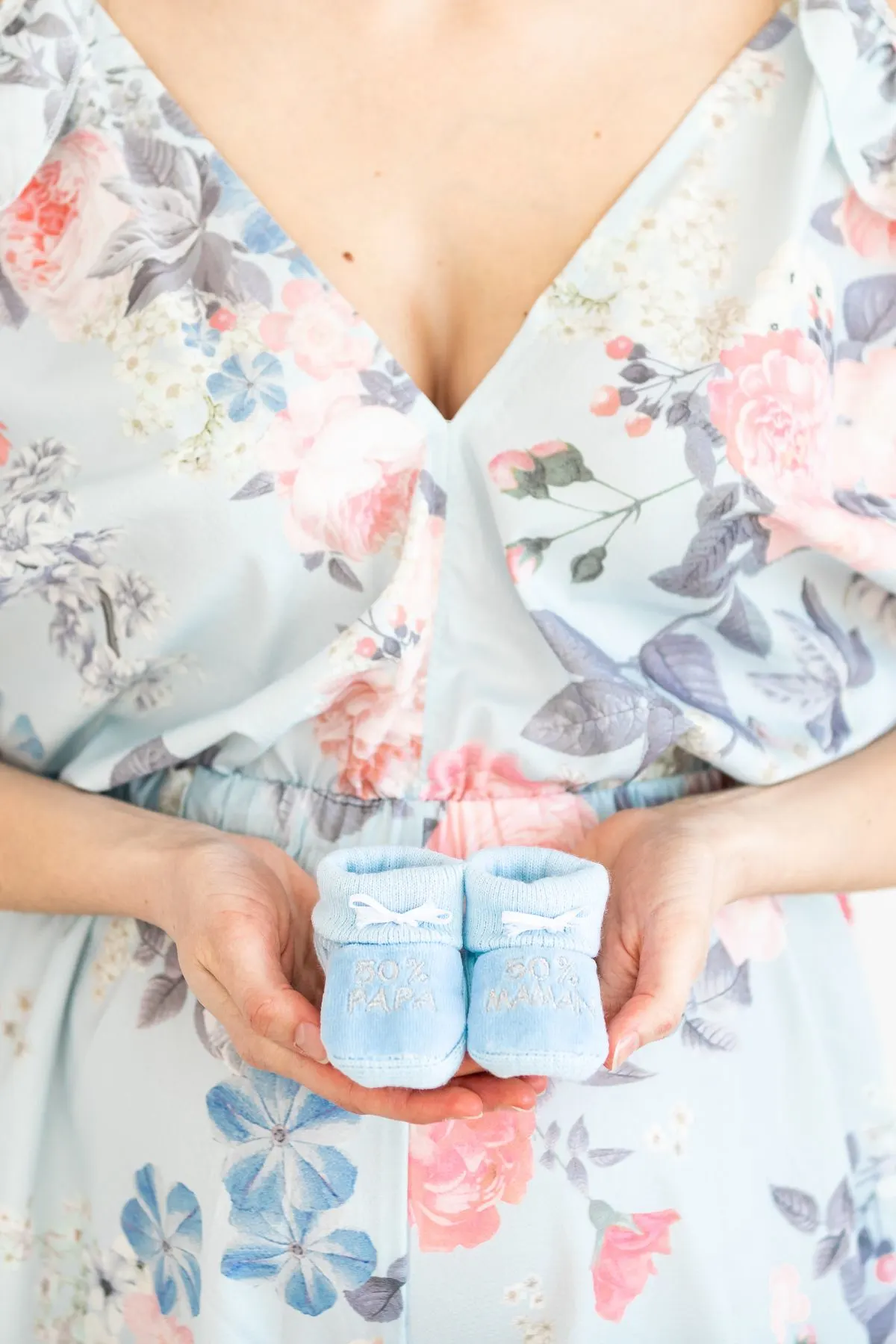 A woman in a pink and blue dress holds blue baby booties in front of her belly.