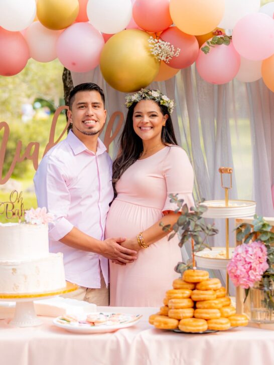 A couple wearing pink poses in front of pink and gold balloons at a baby shower.