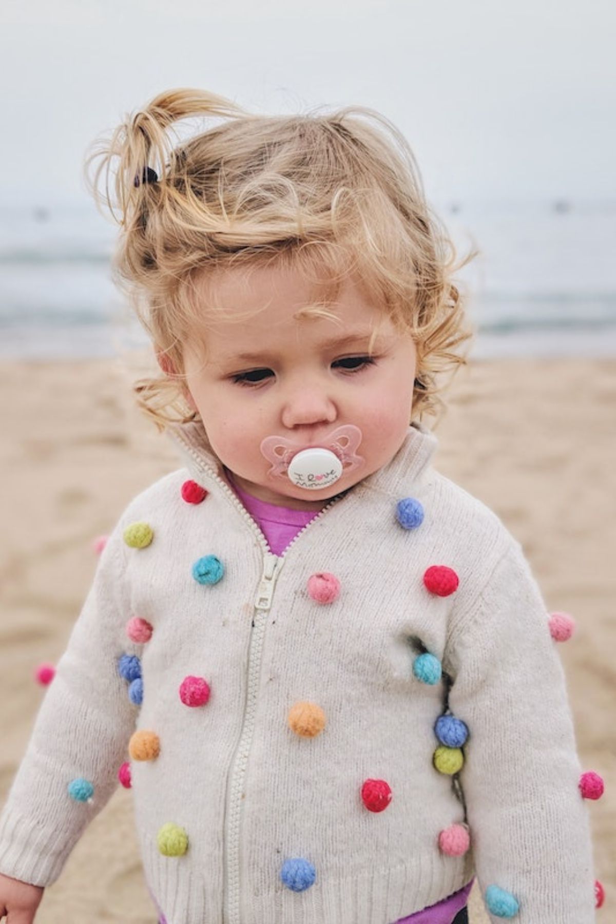 Toddler girl with a pacifier in her mouth walking on the beach.