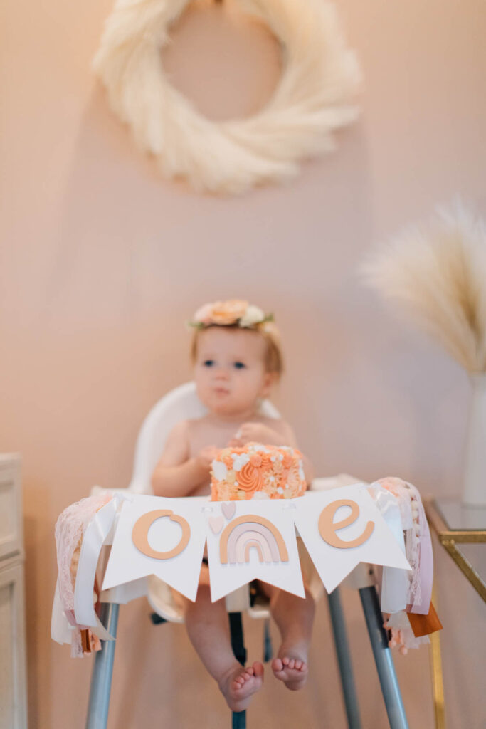 Baby girl eats smash cake in high chair decorated with birthday banner.