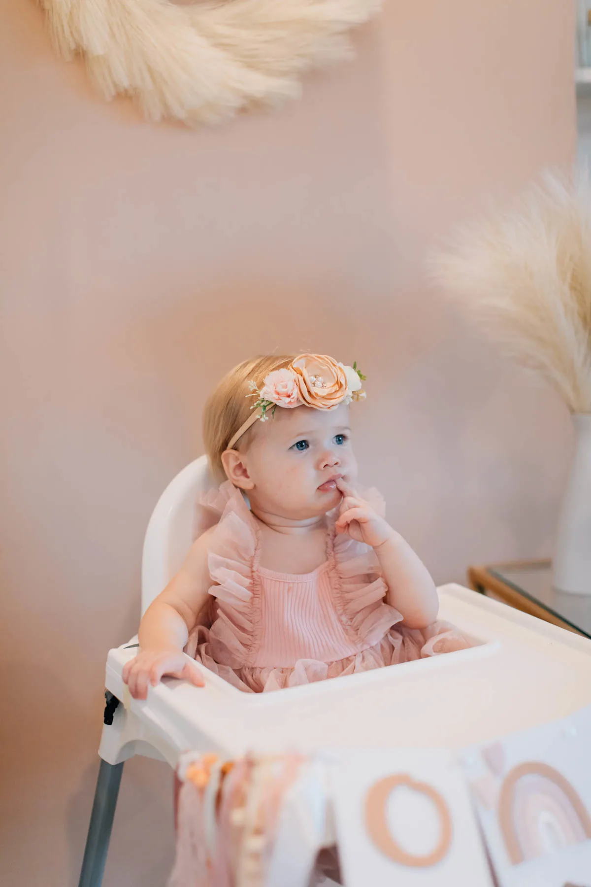 Baby girl wearing pink tulle dress and floral headband sits in white high chair.