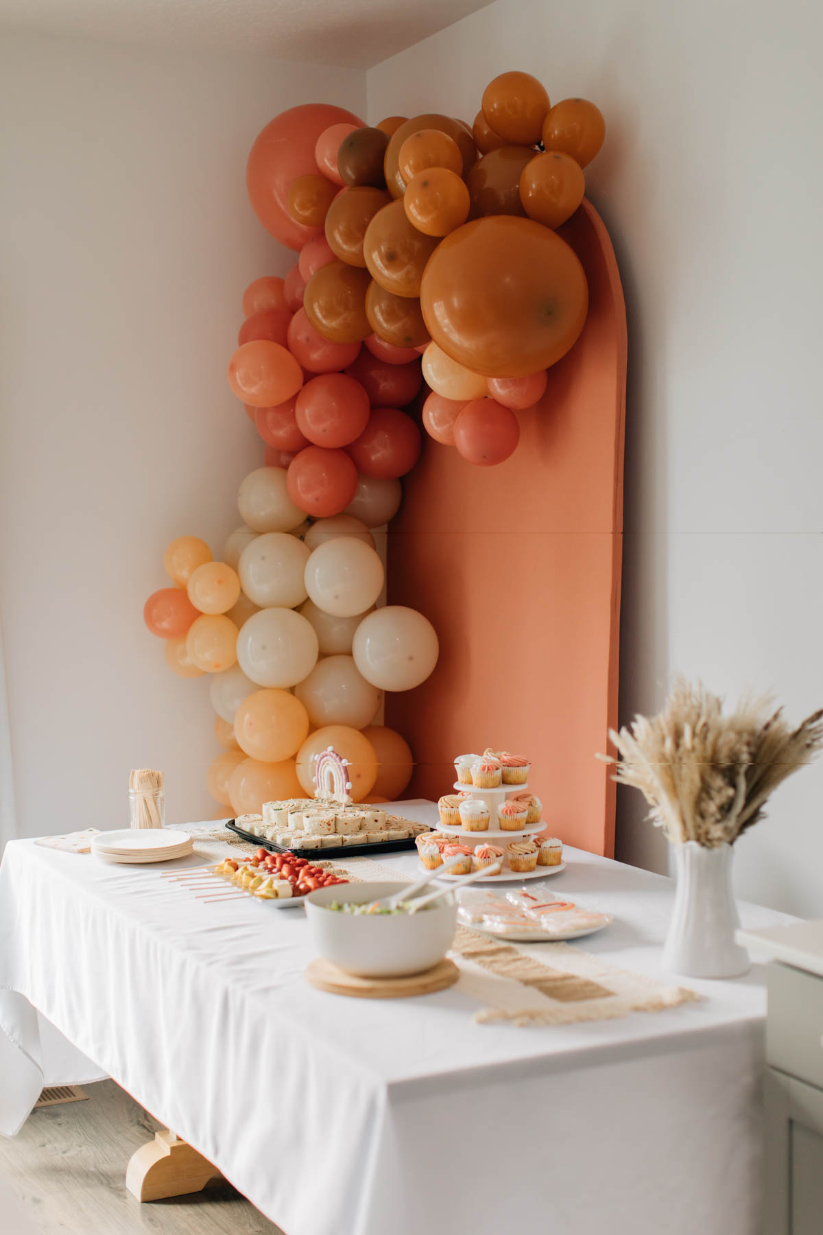 Food table with cupcakes and pampas grass vase in front of arch decor with balloon garland.