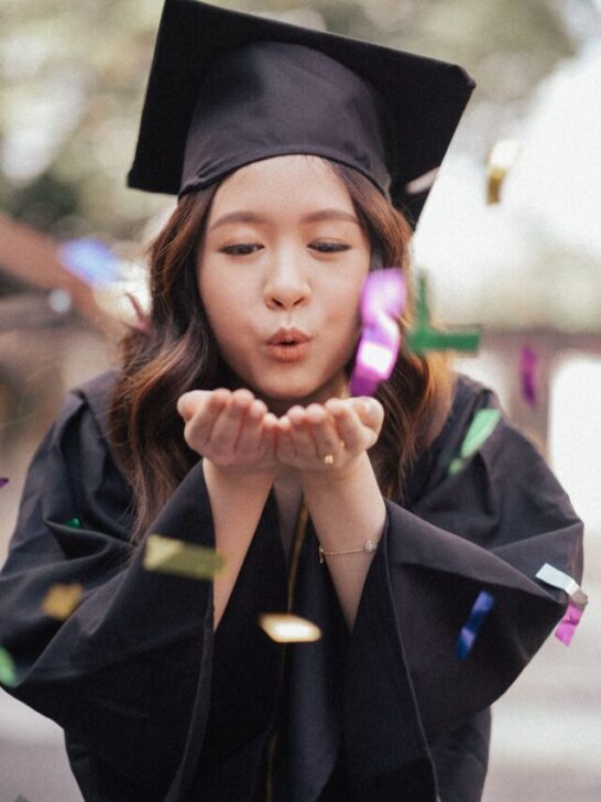 Girl in black graduation cap and gown blowing confetti towards the camera.