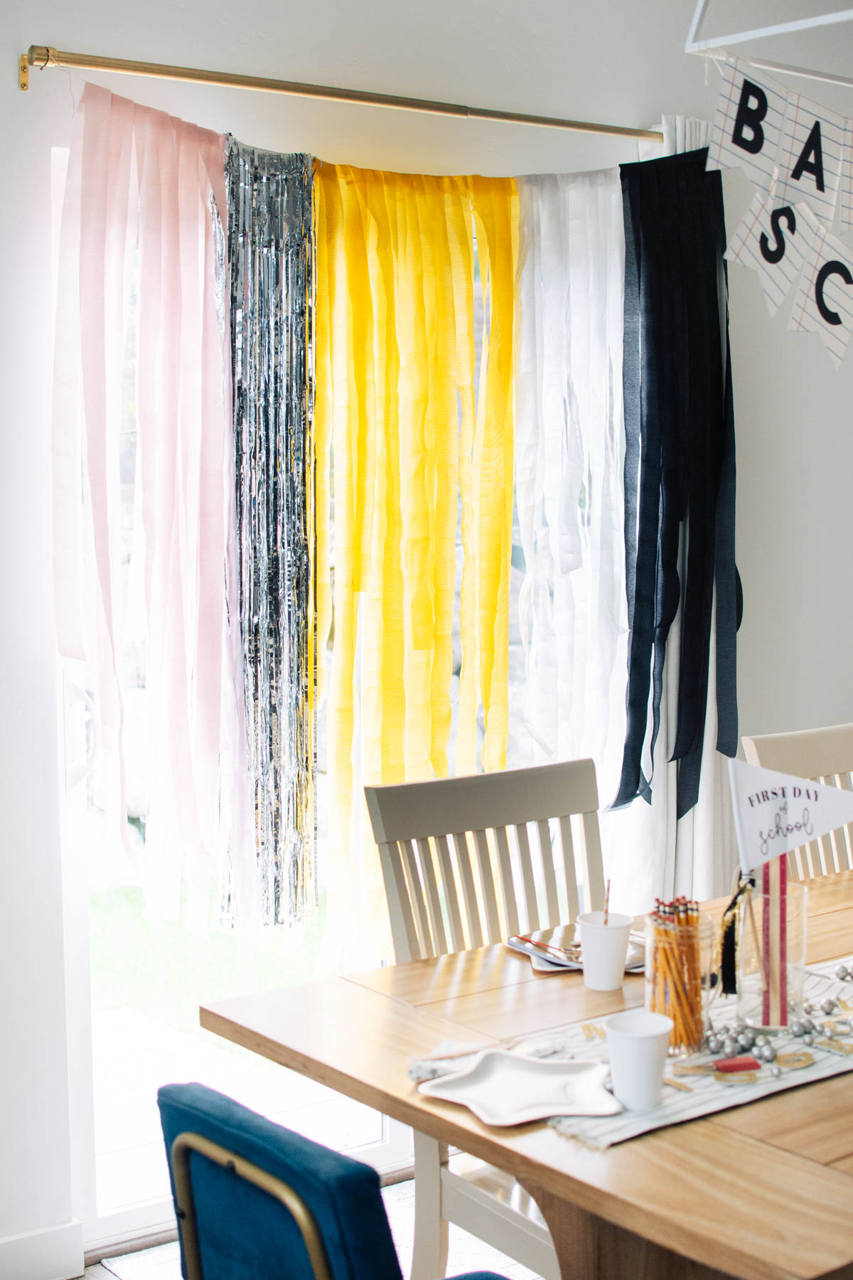 Back to school backdrop with pink, silver, yellow, white, and black streamers hangs from curtain rod behind table.