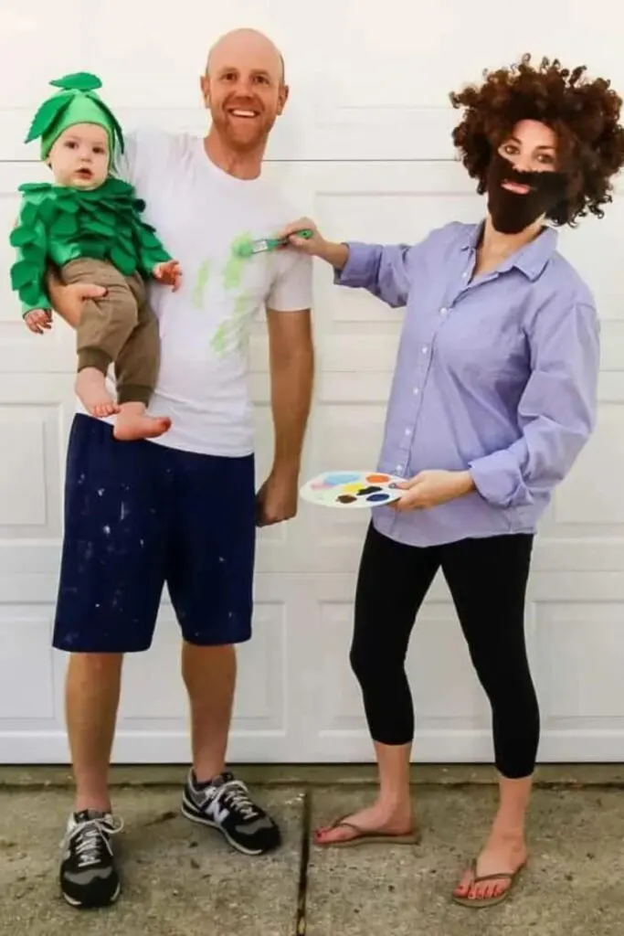 Woman dressed as Bob Ross paints man wearing a white shirt and holding a baby dressed as a tree.