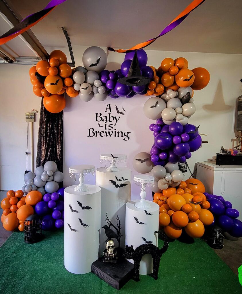 A white backdrop with the phrase "A Baby is Brewing" and gray, orange, and purple Halloween balloons.