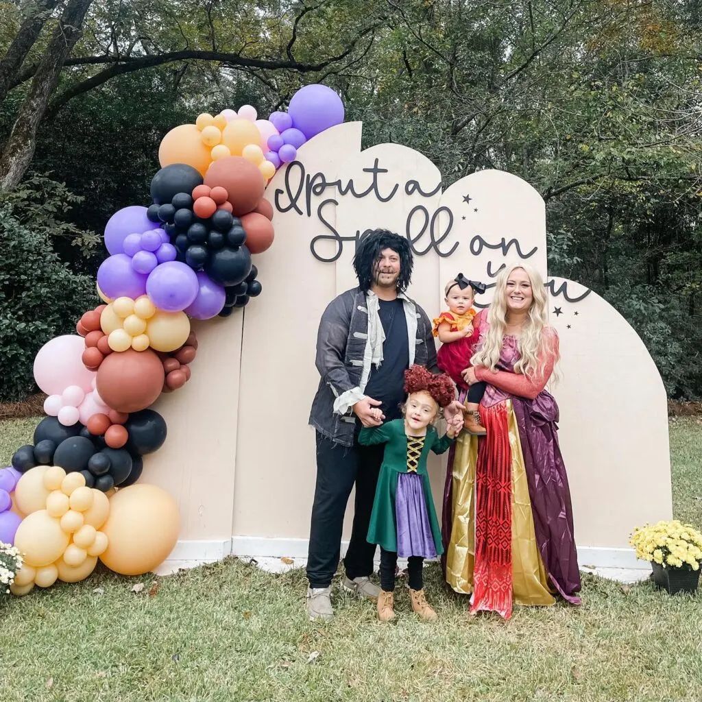 A family dressed in Hocus Pocus Halloween costumes in front of a gender reveal backdrop.