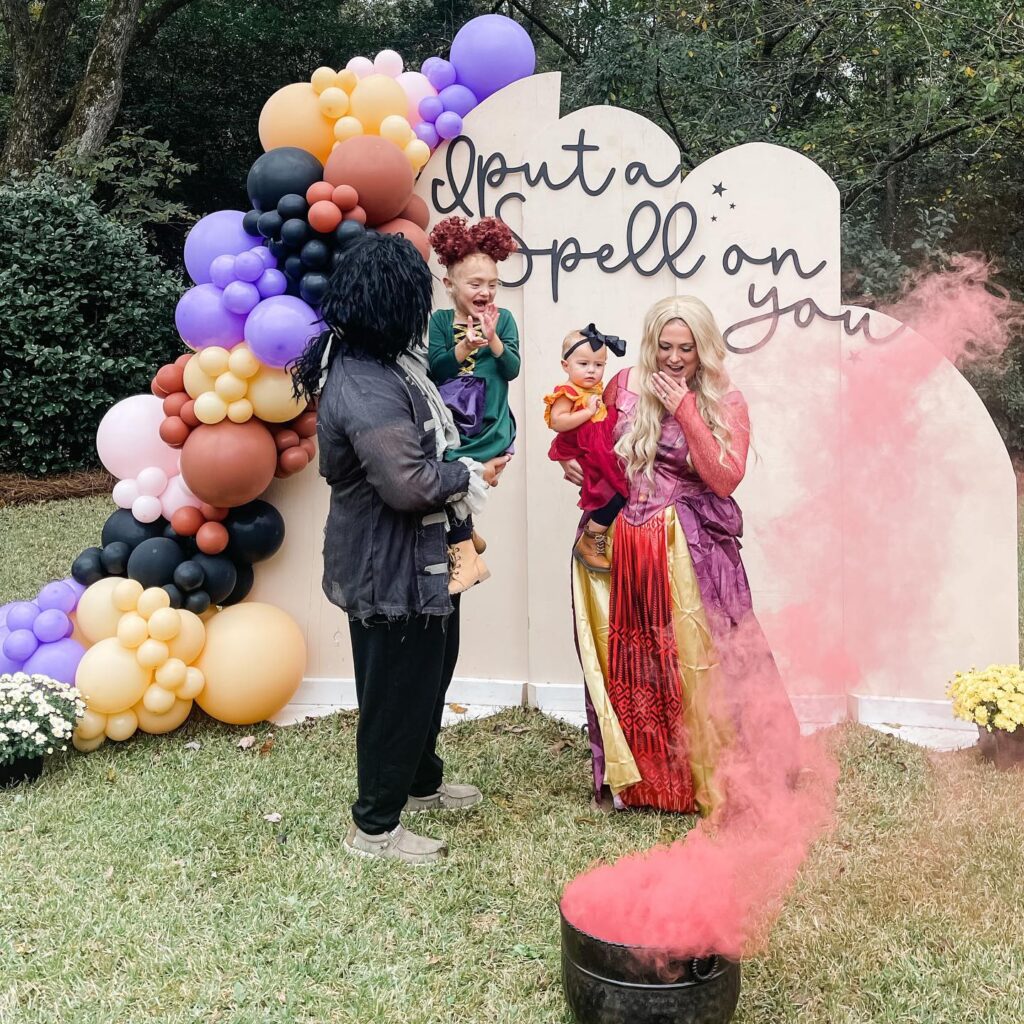 A family in Hocus Pocus costumes stands in front of a back drop with balloons and watching pink smoke rice from a black cauldron.