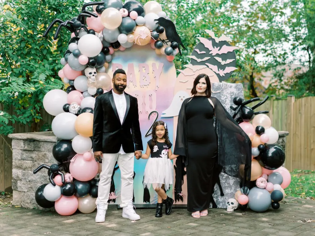 A family stands in front of a pink backdrop with black, white, and pink balloons.
