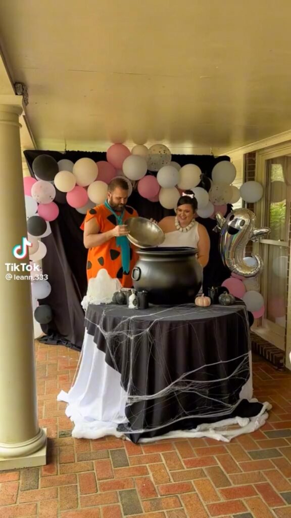 A couple in Flintstone's costumes stands in front of a pink, white, and black balloon arch and looks at a black cauldron.