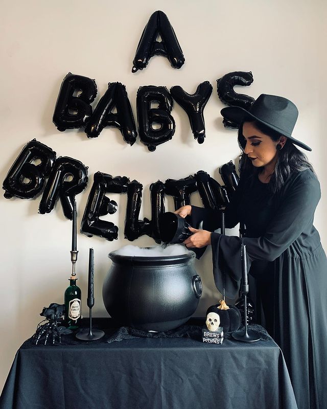 Balloon letters spell "A Baby is Brewing" and a woman dressed as a witch pours smoke into a cauldron.