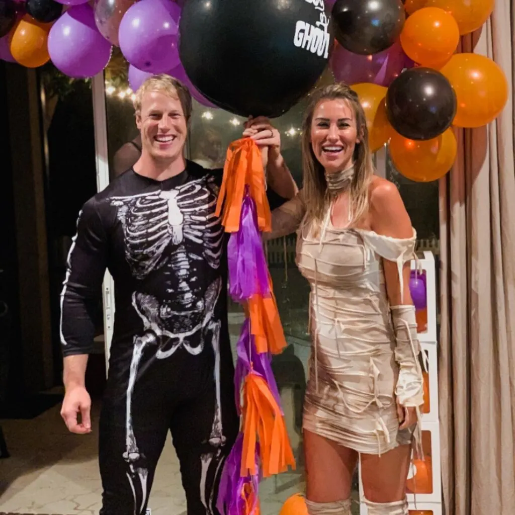 A couple dressed as a skeleton and a mummy hold a black balloon with an orange and purple tail.
