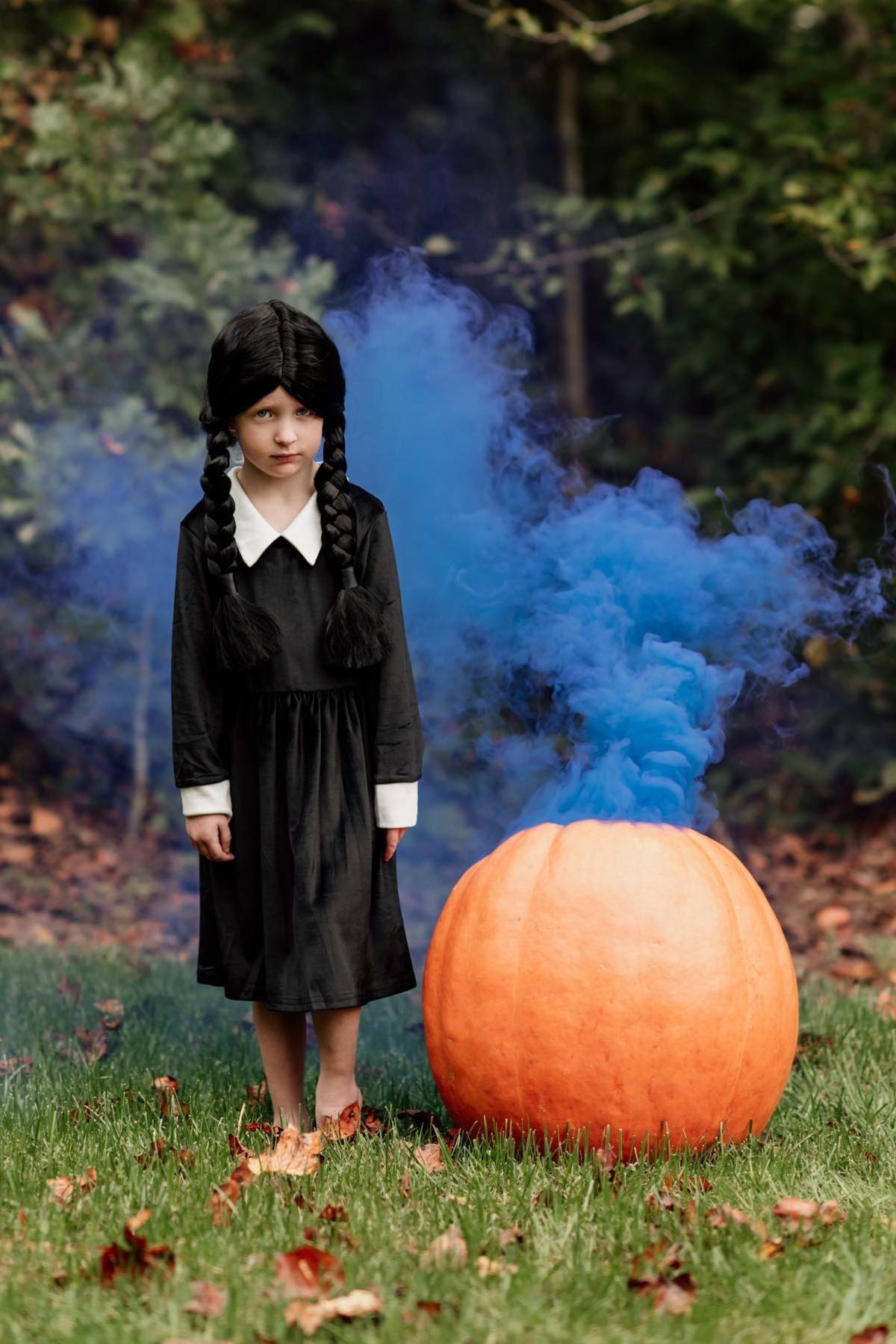 A girl dressed as Wednesday Adams from the Adam's Family next to a pumpkin with blue smoke coming out of it.