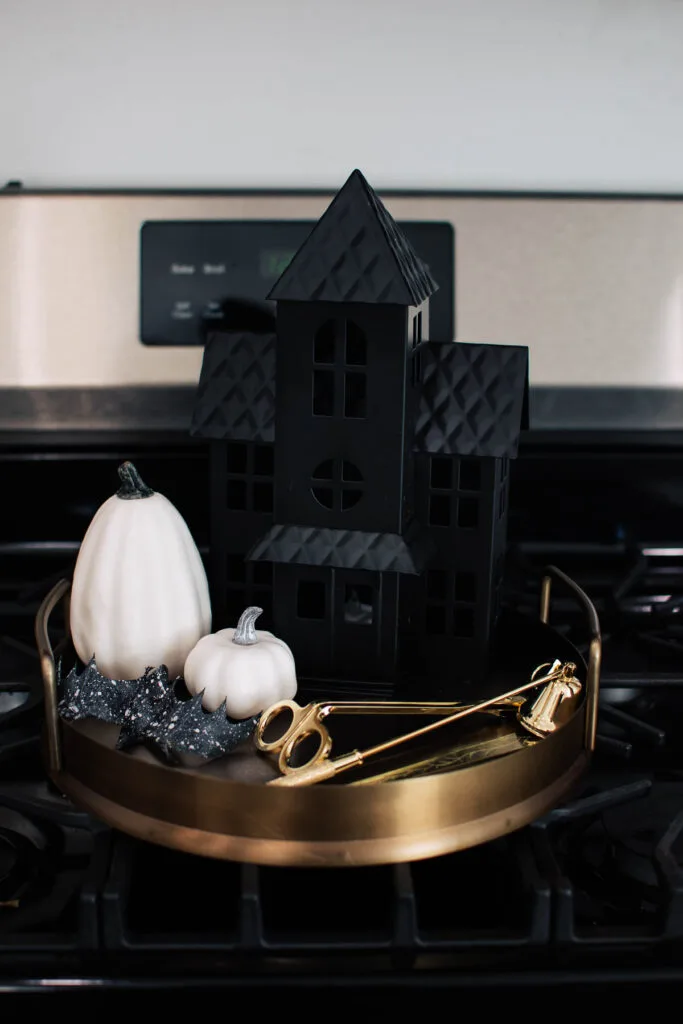 Gold tray with white pumpkins, bat, black haunted house and candle accessories on stove.