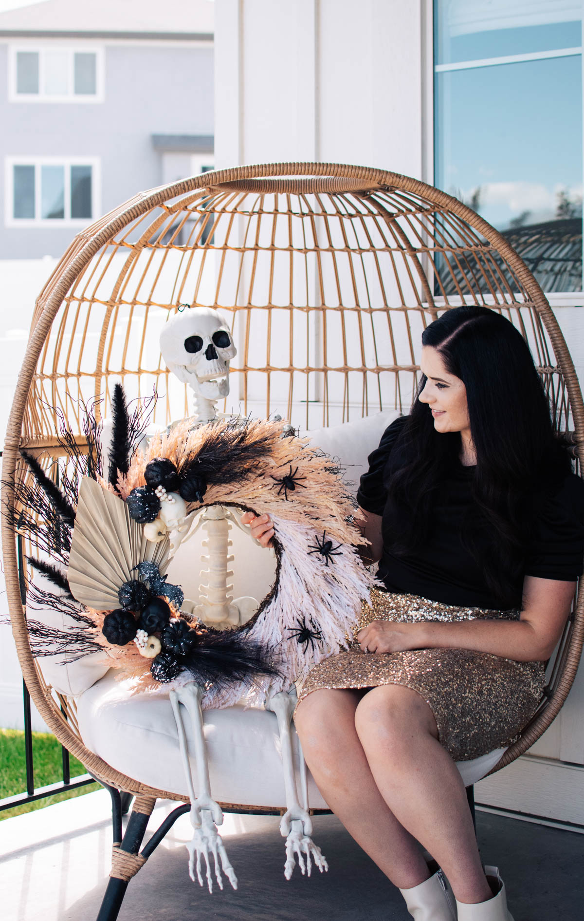 Brunette woman wearing black and gold holds Halloween wreath sitting on skeleton's lap.