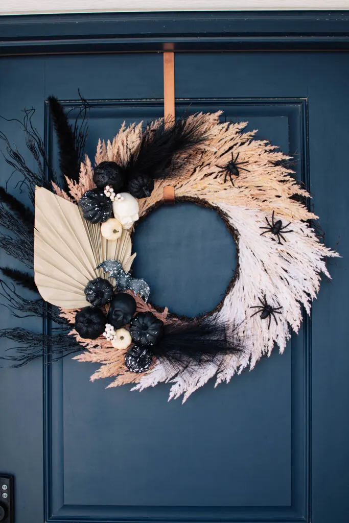 Boho Halloween wreath with pampas grass, skeletons, and pumpkins hangs on navy door.