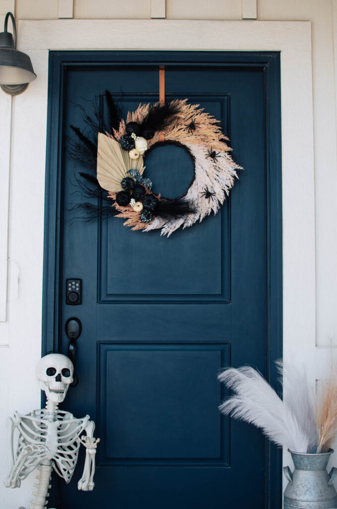 Halloween wreath hangs on navy front door with skeleton and barrel of pampas grass nearby.
