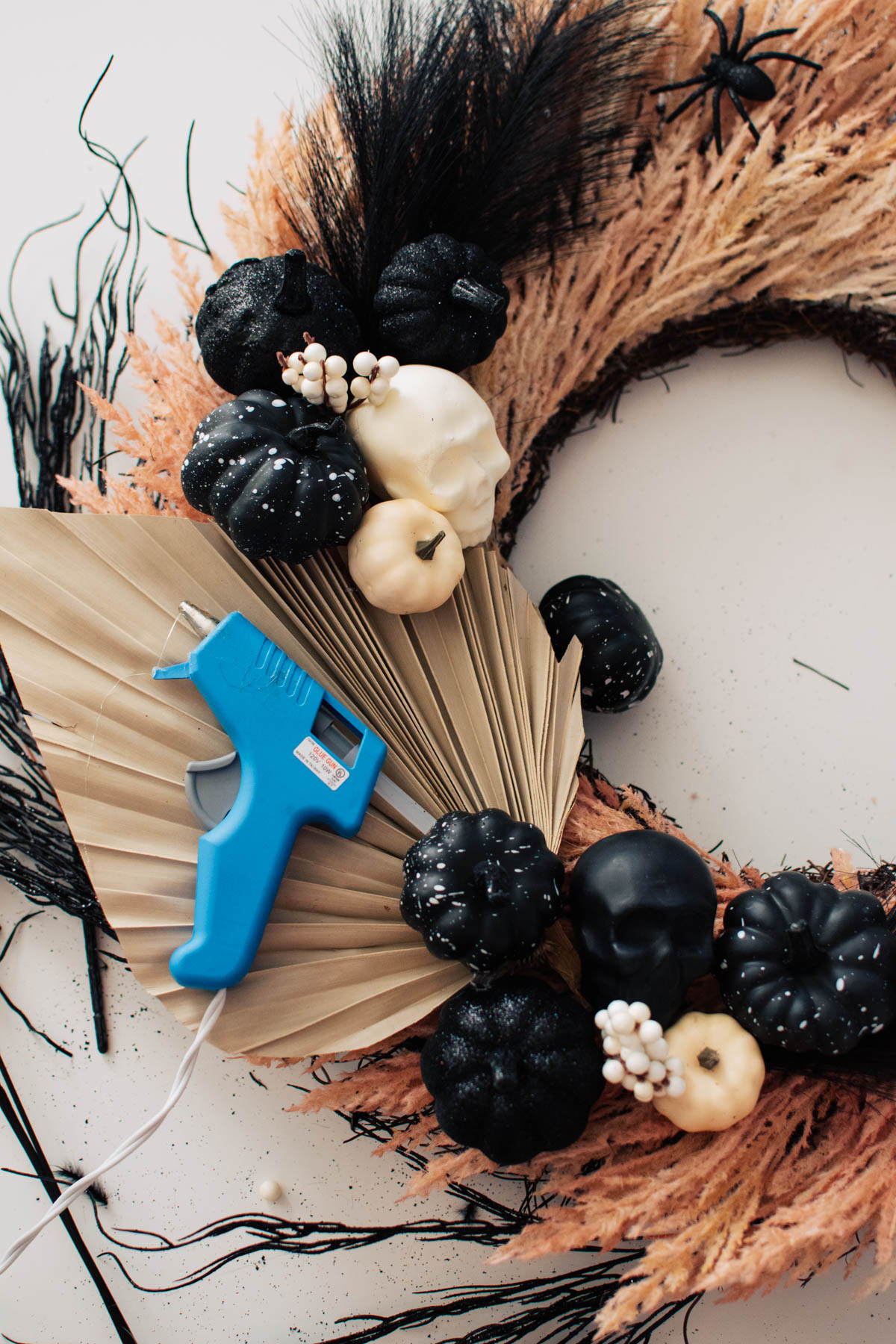 Hot glue gun rests on dried palm leaf next to black and white Halloween items on wreath.