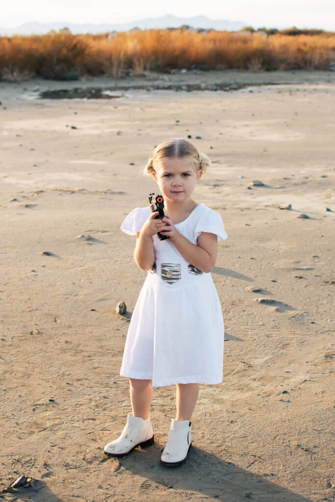 Girl wearing Princess Leia dress holds toy gun in outdoor beach location.