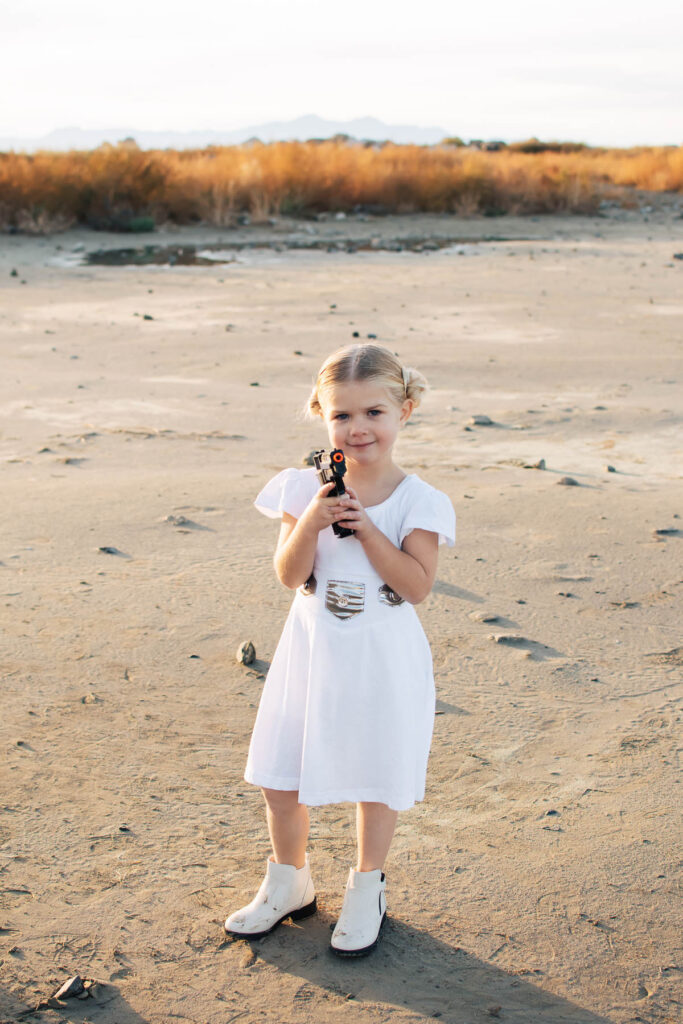Blonde toddler girl wearing Princess Leia dress holds toy gun in outdoor beach location.