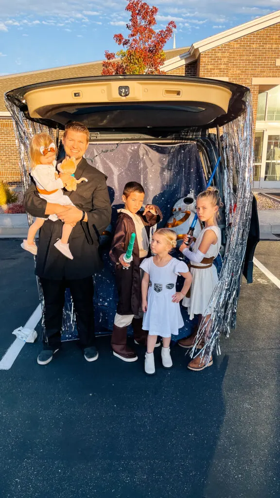 Family dressed as Star Wars characters stands in front of open trunk with decorations.