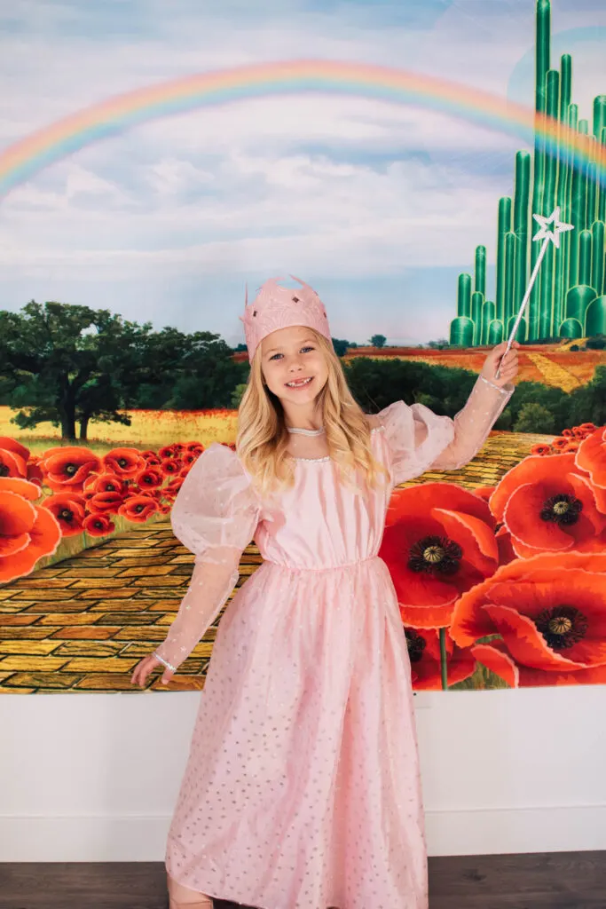 Blonde girl dressed as Glinda stands in front of Emerald City backdrop.