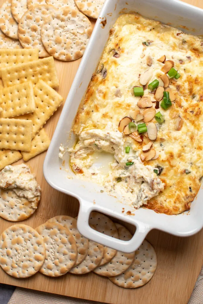 White baking dish of swiss cheese dip with various crackers nearby.