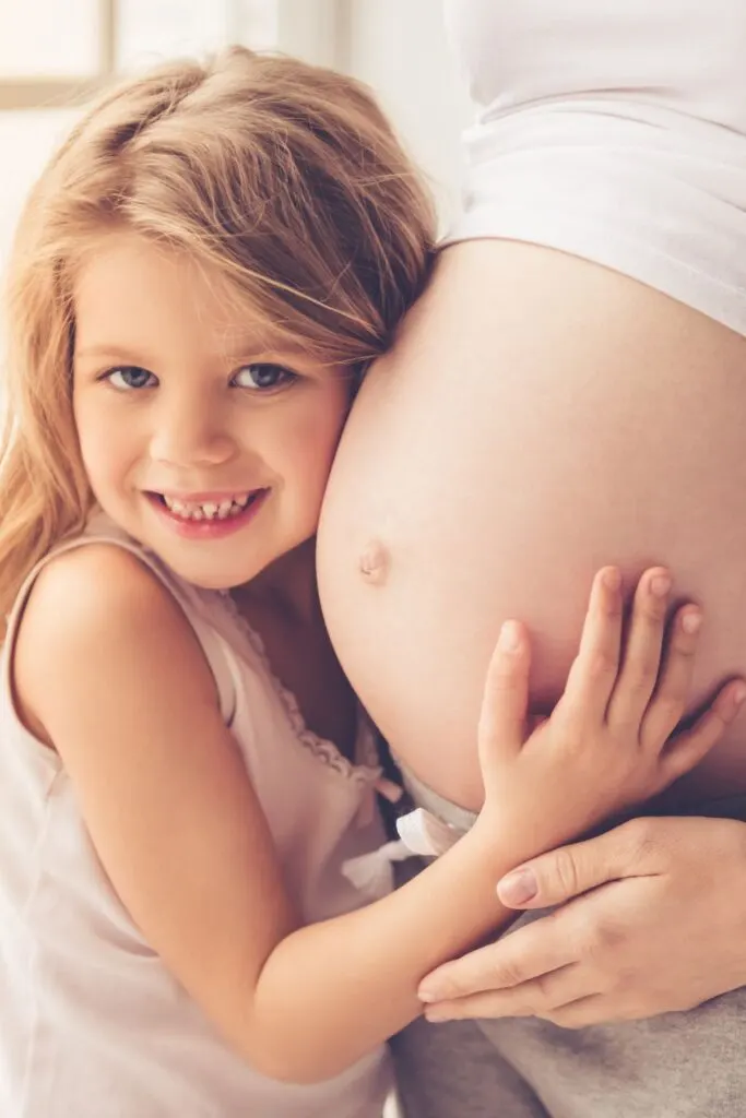 A young girl snuggles up to her pregnant mother's belly while smiling.