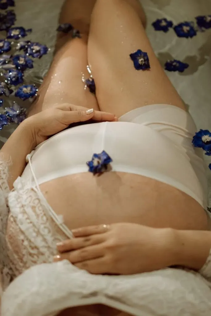 A pregnant woman poses in a milk bath in sheer white under garments with blue flowers.