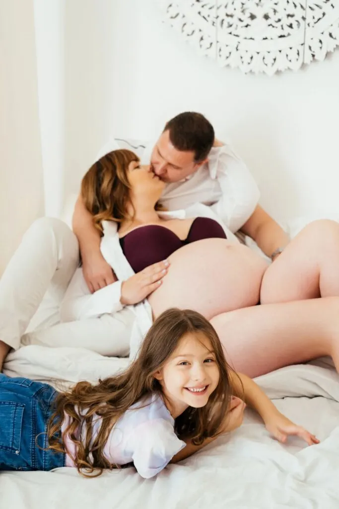 A pregnant woman and man embrace for a picture with their daughter on a bed.